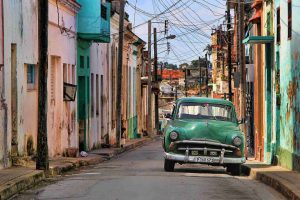 Cuba. Fotografia di una strada che mostra edifici vecchi e fatiscenti su entrambi i lati della strada e un'auto d'epoca verde.