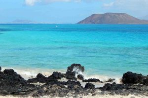 Fuerteventura. Immagine di una spiaggia di sabbia bianca con rocce nere e vista dell'isola Graciosa sullo sfondo.