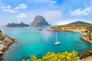Isole Baleari. Fotografia di una baia con acque turchesi e una barca a vela.