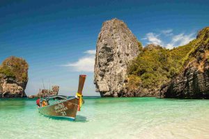 Tailandia. Vista su una spiaggia di sabbia bianca e acque trasparenti con formazioni rocciose sullo sfondo e una barca di legno.