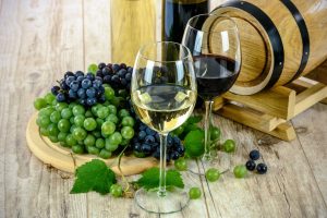 Foto di due bicchieri di vino. Uno di vino rosso e uno di vino bianco. Si possono vedere anche alcuni grappoli d'uva (bianchi e neri). Sullo sfondo due bottiglie di vino e una piccola botte di legno