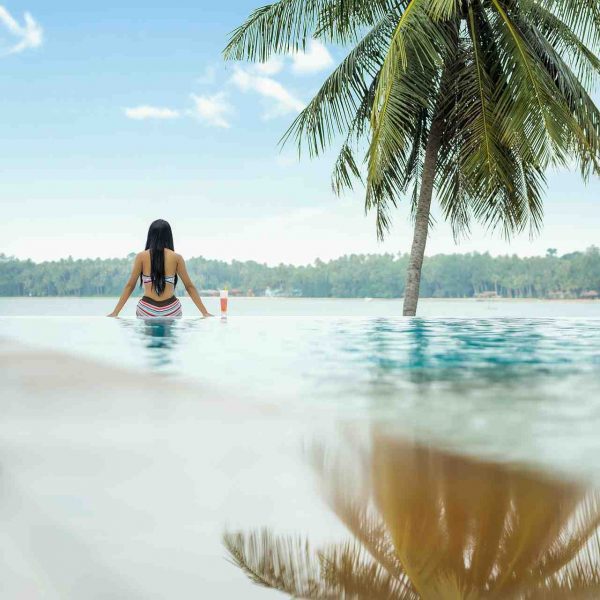 Foto di una ragazza vista di spalle seduta sul bordo di una piscina affacciata su una spiaggia dalla vegetazione lussureggiante